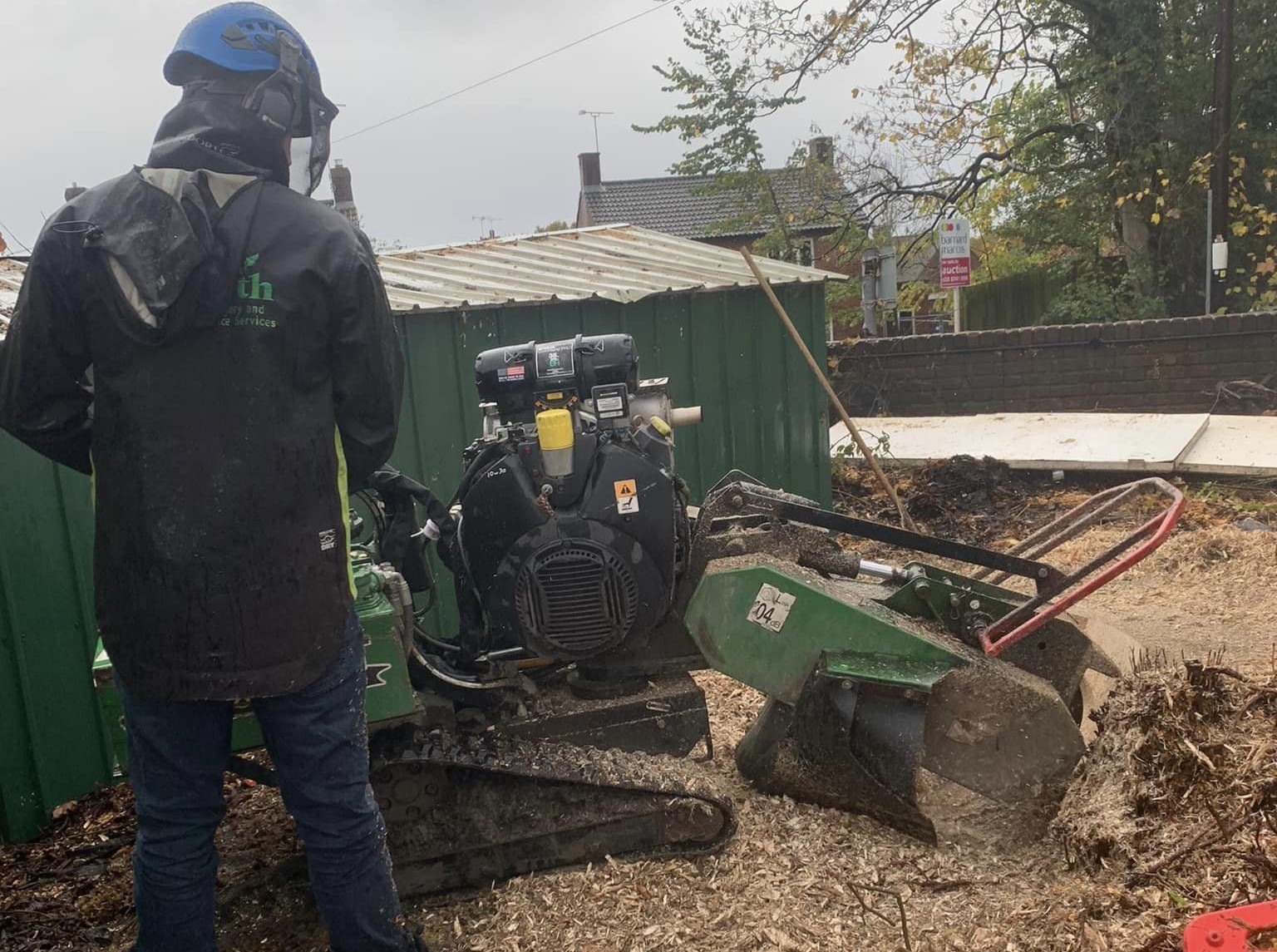 A man in a Greengrowth jacket and protective gear uses a sump grinder to remove a tree stump at ground level.